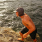 Man standing on a rocky outcrop by the ocean wearing a cap and shorts.