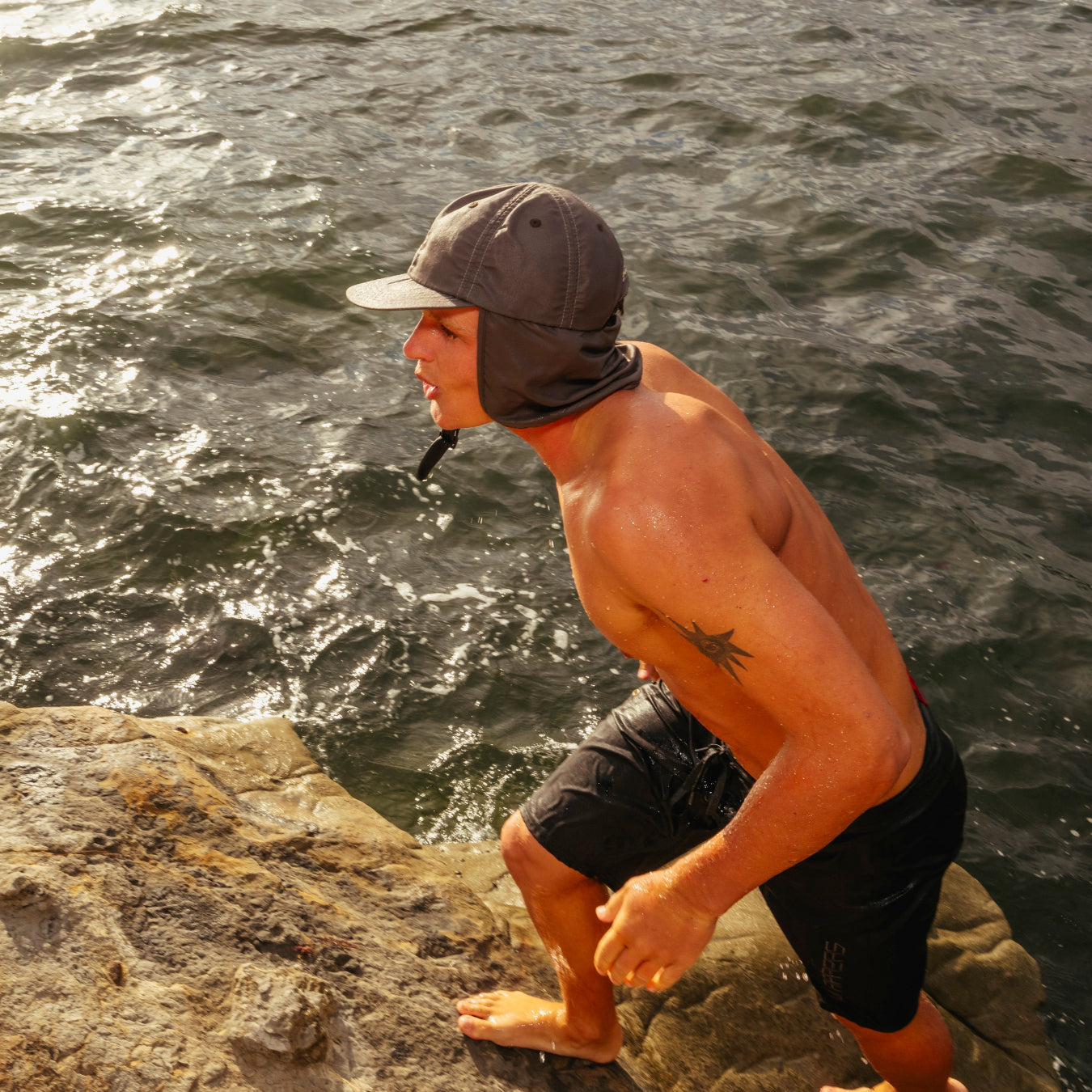 Man standing on a rocky outcrop by the ocean wearing a cap and shorts.